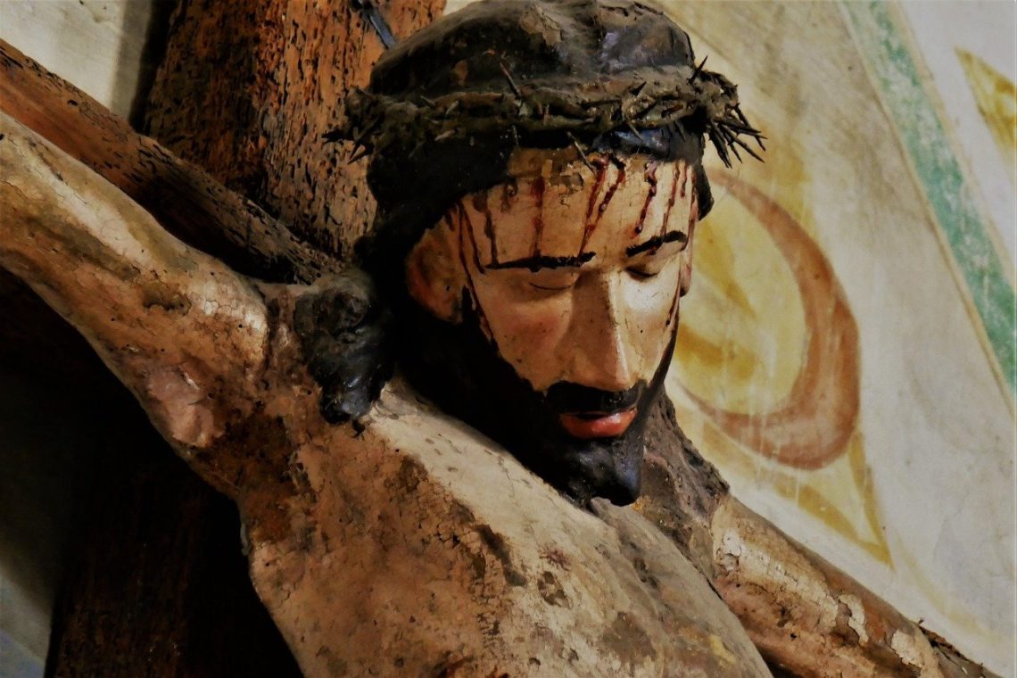 Jezus miłosierny zakrwawiony na drewnianym krzyżu, za nim majaczy fresk świątyni.