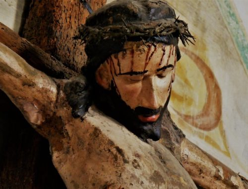 Jezus miłosierny zakrwawiony na drewnianym krzyżu, za nim majaczy fresk świątyni.