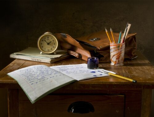 Drewniane biurko, na nim budzik, zeszyty, książki, kredki w kubeczku.