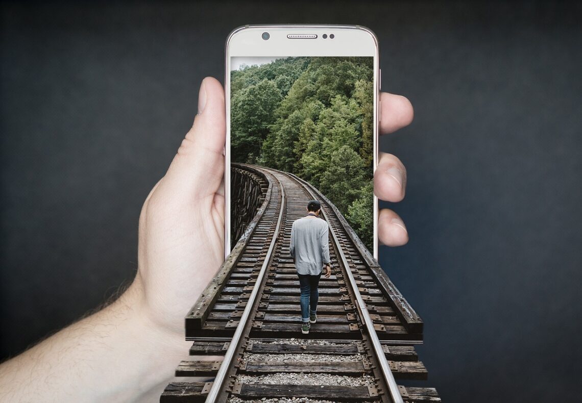 Smartphone w ręce, na telefonie tło z torami w lesie, do telefonu po torach wchodzi człowiek