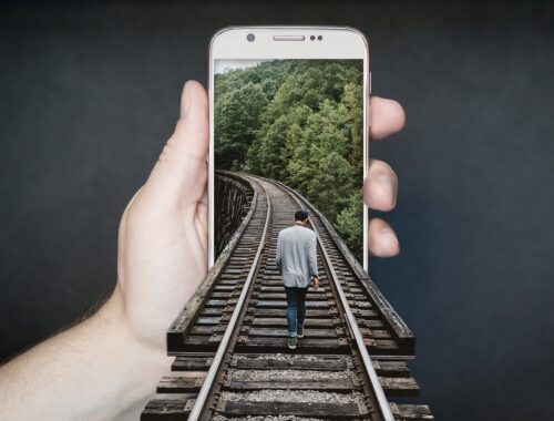 Smartphone w ręce, na telefonie tło z torami w lesie, do telefonu po torach wchodzi człowiek