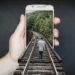 Smartphone w ręce, na telefonie tło z torami w lesie, do telefonu po torach wchodzi człowiek