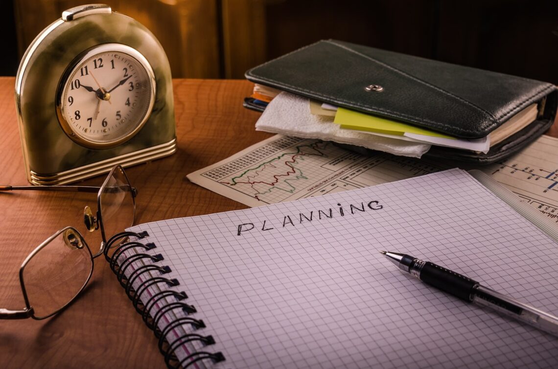 Budzik, notesy, na środku kołonotatnika napis "planning", długopis, okulary ułożone na biurku