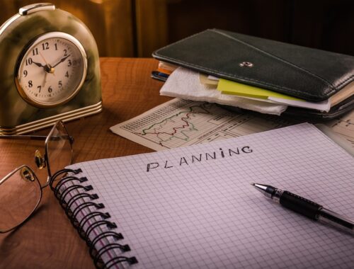 Budzik, notesy, na środku kołonotatnika napis "planning", długopis, okulary ułożone na biurku