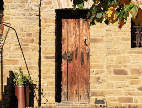 Ceglany dom, drewniane drzwi, doniczka z kwiatami, kilka liści stojącego przed domem dębu, ciepłe światło