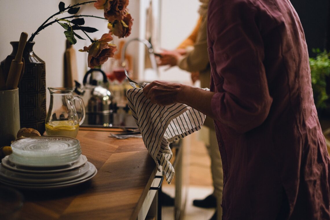 Drewniany stolik, na nim kwiatki, talerze, nad nim ręce kobiety czyszczącej sztuce szmatką materiałową. Wszystko w kolorach retro.